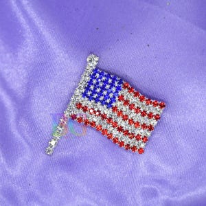 May include: A rhinestone-studded American flag pin. The flag features red and white stripes, a blue field with white stars, and a silver-colored pole. The pin is set against a purple background.