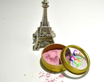 May include: A small, silver Eiffel Tower figurine stands near an open, gold-colored tin filled with pink powder. The tin's lid is partially open, revealing a clear top with colorful text. Pink powder is scattered on the white surface.
