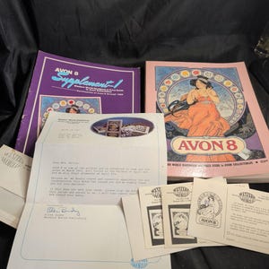 Könnte beinhalten: Zwei Avon 8 Sammlerbücher mit violetten und rosafarbenen Einbänden, die eine Illustration einer Frau in einem fließenden orangefarbenen Kleid zeigen. Enthalten sind ein getippter Brief von Western World Publishing vom 14. März 1987 und kleine Karten.
