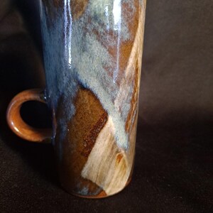 Puede incluir: Una taza cilíndrica alta de cerámica con un esmalte brillante en tonos tierra mezclados de marrón, beige y azul apagado. La taza tiene una superficie lisa y reflectante, y un asa curva unida al lateral con un patrón de colores variado.