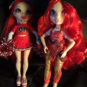 Puede incluir: Dos muñecas con cabello largo rojo y grandes ojos morados. La muñeca de la izquierda viste un conjunto de animadora rojo con el número 86 y sostiene pompones rojos y blancos. La de la derecha lleva una camiseta blanca, chaleco y pantalones rojos.