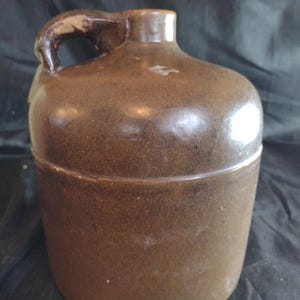 May include: A brown ceramic jug with a cylindrical body, a rounded shoulder, and a short, narrow neck. The jug features a single curved handle attached to the side of the neck and the shoulder. The surface has a smooth, glazed finish.