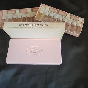 Peut inclure: Deux kits Avon Makeup Demonstrator contenant chacun douze petits flacons en verre de maquillage liquide. Les kits comprennent un boîtier rectangulaire rose clair avec le texte en relief AVON MAKEUP DEMONSTRATOR et une boîte en carton blanche.