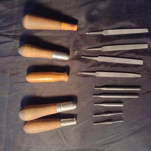 May include: A collection of five hand tools featuring wooden handles and metal ferrules, paired with nine distinct metal file and punch attachments of varying lengths and shapes, arranged in two columns on a dark, textured surface.