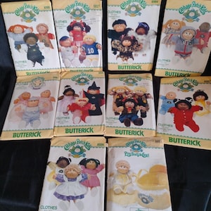 Può includere: Dieci buste di cartamodelli Butterick per Cabbage Patch Kids disposte su tre file. Ogni busta presenta illustrazioni di bambole che indossano vari abiti, il logo Cabbage Patch Kids, i numeri dei modelli e il marchio Butterick in testo verde.