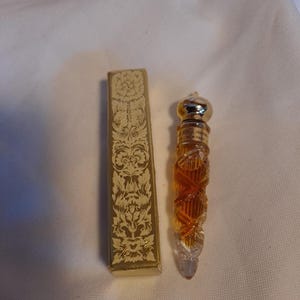 Op de afbeelding: Een hoge, slanke glazen parfumfles met een gefacetteerd amberkleurig lichaam en een goudkleurige dop, naast een rechthoekige goudkleurige doos met een ingewikkeld, in reliëf gemaakt bloemmotief en de tekst Old Rose Perfume New York.