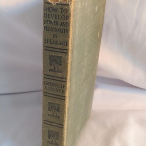 Può includere: Un libro vintage con copertina rigida in tessuto verde chiaro. Il dorso riporta il titolo "HOW TO DEVELOP POWER AND PERSONALITY IN SPEAKING" e l'autore "GRENVILLE KLEISER" in nero, insieme a tre illustrazioni di alberi e un logo.