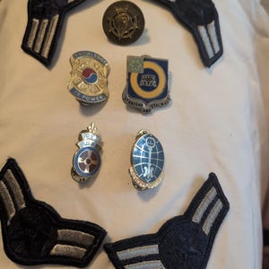 May include: A collection of four military-style pins and four embroidered chevron patches. The pins feature various emblems and text including Knowledge Is Power and Straight And Stalwart. The patches are dark blue with silver-toned stripes.