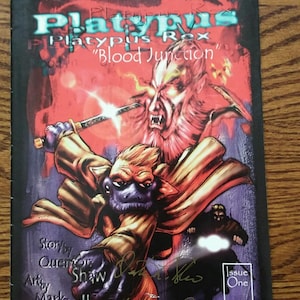 May include: A comic book titled Platypus Rex Blood Junction, Issue One, featuring a central character in a tan trench coat holding a sword, with a red-toned monstrous figure behind it. The cover includes text credits for Quenton Shaw and Mark Brill.