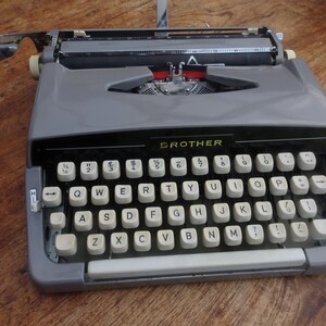 May include: A gray Brother typewriter with a black keyboard and white keys. The keys have letters, numbers, and symbols on them. The typewriter has a carriage return lever and a paper release lever.