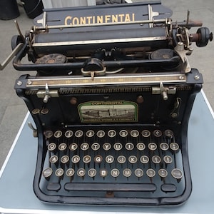 May include: A black vintage typewriter with a gold-colored keyboard and a label that reads "CONTINENTAL Wanderer Werke A.G. Chemnitz".