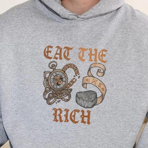 May include: A light gray hoodie with a drawstring hood. The front features the phrase "EAT THE RICH" in a copper-colored, ornate font, along with a pocket watch, scroll, and a rock graphic.