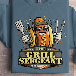 May include: Blue sweatshirt featuring a cartoon hot dog character wearing sunglasses and a headband, holding a spatula and tongs. The text "THE GRILL SERGEANT" is displayed below the character in a banner.