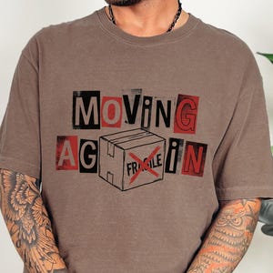 May include: Brown t-shirt with the words "MOVING AGAIN" in a cut-out style, with a box graphic and the word "FRAGILE" crossed out. The shirt is a neutral color and has short sleeves.
