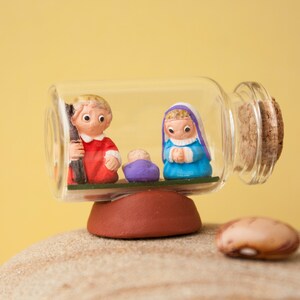 May include: A clear glass bottle with a cork top containing a miniature nativity scene. The scene includes a red-clad Joseph, a blue-clad Mary, and a purple-clad baby Jesus. The bottle is sitting on a brown rock.