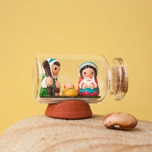 May include: A miniature nativity scene inside a clear glass bottle with a cork stopper. The figures are painted with bright colors and sit on a small, red base. A single bean rests on a stone in the foreground.