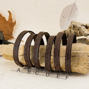 May include: Six brown leather bracelets with an embossed floral design. The bracelets are arranged in a row on a piece of driftwood.