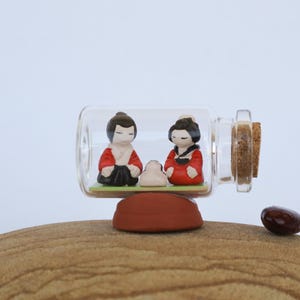 May include: A glass bottle with a cork stopper contains two small ceramic figurines of people wearing red and black clothing. The bottle is sitting on a brown rock.