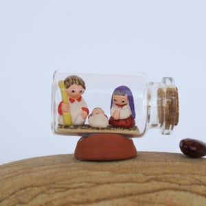 May include: A miniature nativity scene inside a clear glass bottle with a cork stopper. The scene features figurines of Mary, Joseph, and baby Jesus. The bottle rests on a brown, rounded base.
