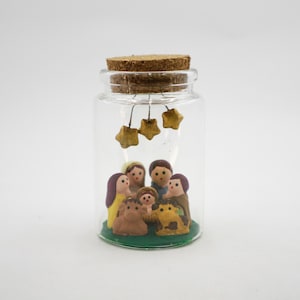May include: A clear glass jar with a cork stopper, containing a miniature nativity scene. The scene includes small figurines of people and animals, with hanging star decorations. The base is green.