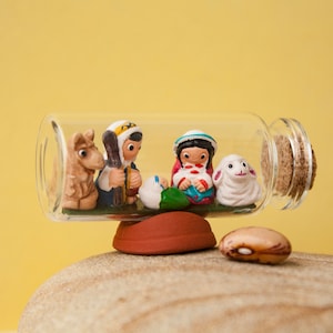 May include: A miniature nativity scene inside a clear glass bottle with a cork stopper. The scene includes figurines of a camel, shepherd, Mary, baby Jesus, and a lamb. The bottle rests on a small, round, red-brown base.