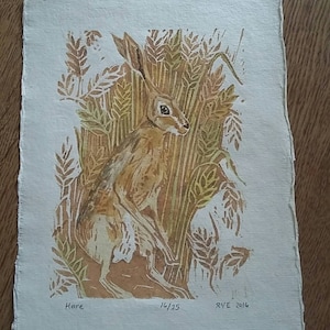 Op de afbeelding: Een bruine haasprint op gestructureerd papier met een achtergrond van een tarweveld. De print is genummerd 16/25 en gesigneerd "RYE 2016".