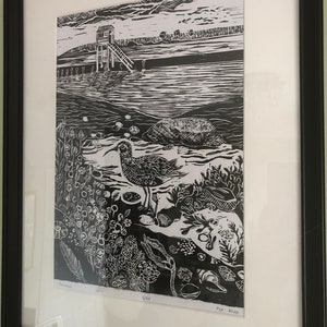 May include: Framed monochrome print of a coastal landscape. The artwork shows a bird in the foreground, a pier, and a small building. The print is signed and numbered, with the year 2020 visible.