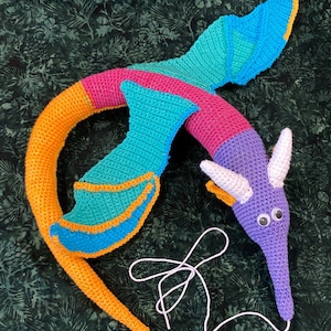 Puede incluir: Un juguete de gusano de ganchillo hecho a mano y colorido. El gusano presenta segmentos de color naranja, rosa, turquesa, morado y azul. Tiene cuernos blancos, ojos saltones y un cuerpo largo y delgado con aletas. Una cuerda blanca está adjunta.