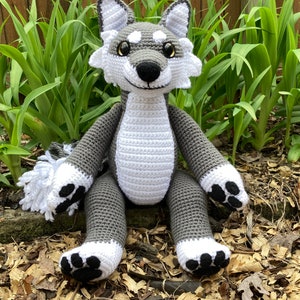 Op de afbeelding: Een gehaakt grijs en wit wolvenknuffel met zwarte ogen en een witte neus. De wolf zit op een bed van bruine houtsnippers.