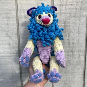 Puede incluir: Muñeca de león de ganchillo hecha a mano con melena azul, orejas moradas y nariz rosa. El león tiene brazos y piernas color crema con detalles morados y azules. La muñeca está sentada, mostrando sus patas.