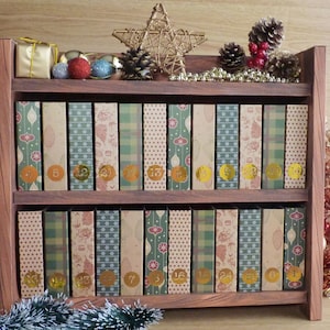 Op de afbeelding: Een houten adventskalender met twee planken, elk met twaalf genummerde dozen met feestelijke patronen. De bovenste plank heeft een gouden geschenkdoos, ornamenten, dennenappels en een ster. De kalender is versierd voor de feestdagen.