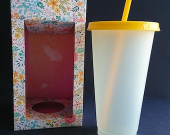 Pode incluir: Um copo branco fosco com tampa amarela e um canudo amarelo. O copo está sobre uma mesa ao lado de uma caixa com um padrão floral.