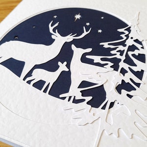 May include: A white paper card with a blue circle cut out in the center. The circle features a white silhouette of a deer family with a star in the sky. The card is decorated with a white paper tree silhouette.