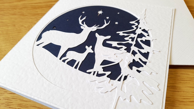 May include: A white paper card with a blue circle cut out in the center. The circle features a white silhouette of a deer family with a star in the sky. The card is decorated with a white paper tree silhouette.