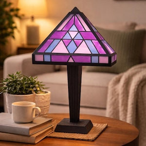 Può includere: Lampada da tavolo decorativa con paralume in vetro colorato nei toni del viola, rosa e blu. La lampada ha una base e uno stelo neri e poggia su un tavolo di legno. Il paralume ha un design geometrico.