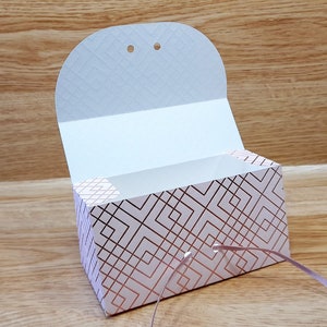 May include: A white gift box with a geometric pattern in rose gold. The box has a flap that opens to reveal the inside. The box is tied with a rose gold ribbon.