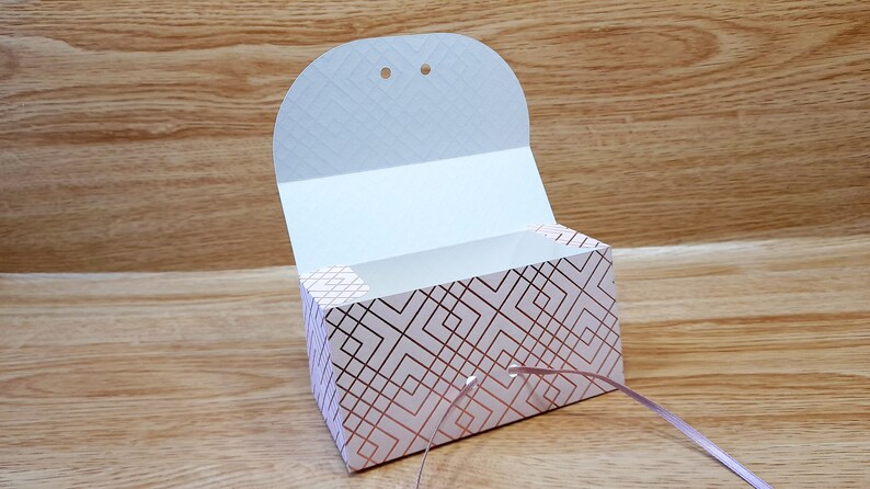 May include: A white gift box with a geometric pattern in rose gold. The box has a flap that opens to reveal the inside. The box is tied with a rose gold ribbon.
