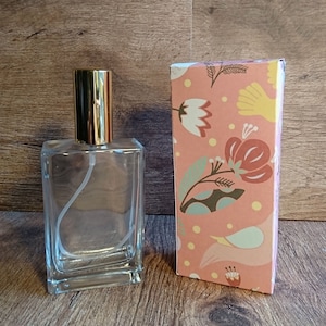 May include: A clear glass bottle with a gold spray nozzle and a matching gold cap. The bottle is empty and is sitting on a wooden surface. Next to the bottle is a pink box with a floral pattern.