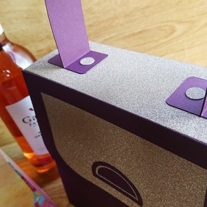Pode incluir: Uma caixa de presente roxa com acabamento de glitter prateado e uma al&ccedil;a roxa. A caixa tem um pequeno ret&acirc;ngulo arredondado roxo na frente. A caixa est&aacute; sobre uma superf&iacute;cie de madeira.