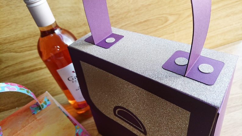 Pode incluir: Uma caixa de presente roxa com acabamento de glitter prateado e uma al&ccedil;a roxa. A caixa tem um pequeno ret&acirc;ngulo arredondado roxo na frente. A caixa est&aacute; sobre uma superf&iacute;cie de madeira.