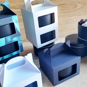 May include: Four gift boxes with windows to display the contents. Two boxes are white, one is black with a green leaf pattern, and one is black. The boxes have handles and are ideal for packaging candles or other small gifts. Three black metal tins with lids are also shown, one containing white and purple heart-shaped candies.
