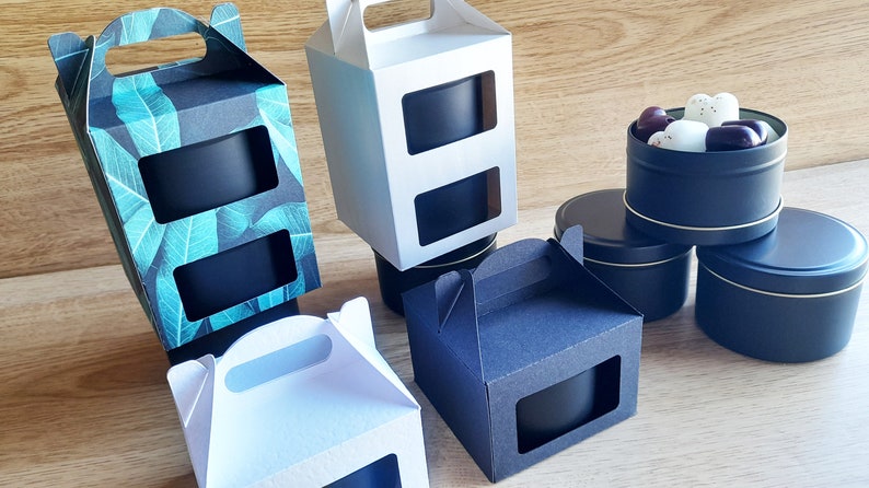 May include: Four gift boxes with windows to display the contents. Two boxes are white, one is black with a green leaf pattern, and one is black. The boxes have handles and are ideal for packaging candles or other small gifts. Three black metal tins with lids are also shown, one containing white and purple heart-shaped candies.