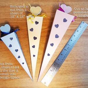 May include: Four paper cones with heart-shaped tops in blue, yellow, pink, and white. The cones are decorated with black hearts and are arranged on a wooden surface with a ruler for scale. The text "Enlarged to cut from a sheet of 12" x 12"" is above the yellow cone. The text "Cut as large as possible from a single sheet of A3" is above the pink cone. The text "Cut from a sheet of A4 at the size it comes into Design Space" is below the blue cone.