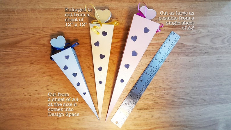 May include: Four paper cones with heart-shaped tops in blue, yellow, pink, and white. The cones are decorated with black hearts and are arranged on a wooden surface with a ruler for scale. The text "Enlarged to cut from a sheet of 12" x 12"" is above the yellow cone. The text "Cut as large as possible from a single sheet of A3" is above the pink cone. The text "Cut from a sheet of A4 at the size it comes into Design Space" is below the blue cone.