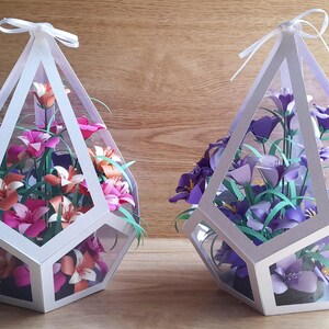 May include: Two white geometric paper flower arrangements. One arrangement features pink and orange flowers, the other features purple flowers. Both arrangements are in a clear geometric vase.