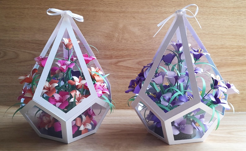 May include: Two white geometric paper flower arrangements. One arrangement features pink and orange flowers, the other features purple flowers. Both arrangements are in a clear geometric vase.