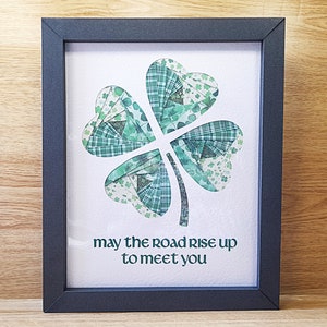 May include: A framed print with a green shamrock made of fabric scraps. The print has the text "may the road rise up to meet you".