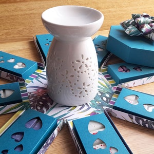 May include: A teal blue gift box with a green and white bow, surrounded by teal blue boxes with heart-shaped cutouts. A white ceramic oil diffuser with a floral design sits in the center.