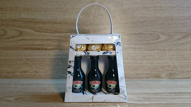 Pode incluir: Uma sacola de presente branca com al&ccedil;a, com um padr&atilde;o de m&aacute;rmore e uma janela transparente. Dentro da sacola, tr&ecirc;s garrafas em miniatura de Baileys Irish Cream e alguns chocolates embrulhados em ouro.