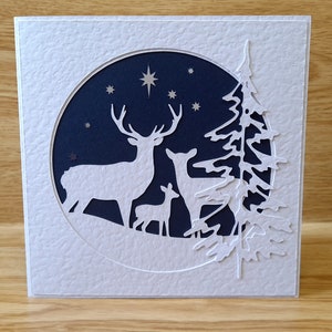 May include: A white card with a blue circle cut out in the center. The circle features a silhouette of three deer in a snowy forest with stars in the night sky. The card is decorated with a white silhouette of a pine tree.